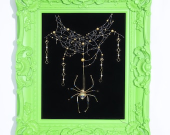 May include: A bright lime green ornate frame containing a black background with a delicate gold wire spider web and a central spider figure. The spider features gold legs and a blue and gold body, with dangling gold chains and small beads.
