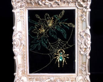 May include: A rectangular ornate silver-toned frame containing a black background with a gold and green wire-art spider web, a central spider with a green gemstone body, and gold and green wire-art lily flowers positioned above the web.