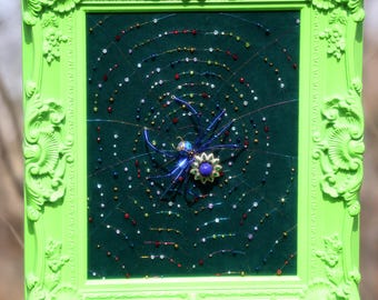 May include: A framed artwork featuring a dark green background with a beaded spiderweb design. The spider is adorned with a blue and purple jewel. The frame is a bright, ornate green.