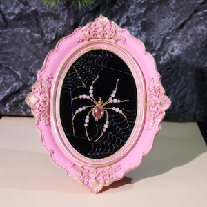 Può includere: Una cornice ovale rosa ornata con un bordo interno dorato circonda uno sfondo nero. Un ragno fatto di filo d'oro e perline rosa è seduto su una ragnatela. La cornice è decorata con accenti floreali, creando un pezzo decorativo.