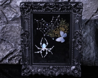 May include: A framed artwork featuring a spider, web, and butterfly design. The black frame surrounds a black background with a white spider hanging from a web. A blue butterfly and gold floral accents complete the design.