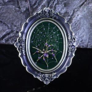 May include: An oval framed artwork featuring a beaded spider and web design. The spider is purple and gold, with a web of gold thread and purple beads. The frame is dark blue with ornate silver detailing, set against a dark green background.