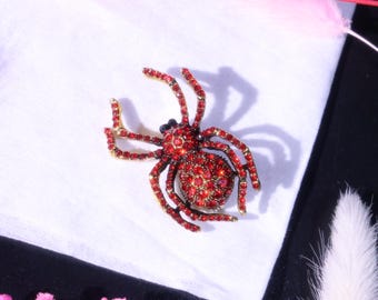 May include: A spider-shaped brooch featuring a gold-tone metal frame encrusted with numerous small, vibrant red rhinestones covering the body and all eight legs. The design includes a rounded abdomen and a smaller head section with dark accents.