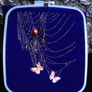 May include: A square embroidery hoop containing a deep blue fabric background. A spider with a bright red abdomen and black legs sits in a web made of thin silver threads adorned with small gold beads. Two butterflies are attached to the web.