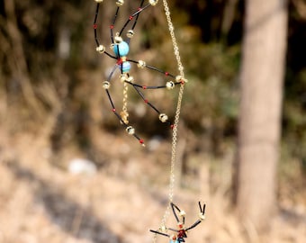 May include: Two decorative spider ornaments hanging from a gold chain. The spiders have black wire bodies, red accents, and gold bead details. Each spider features a turquoise bead as the body center.