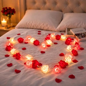 May include: A romantic scene on a bed with a heart-shaped arrangement of red and white rose string lights. Scattered red rose petals enhance the ambiance. A vase of roses and a lit candle are on the nightstand, creating a warm, inviting atmosphere.