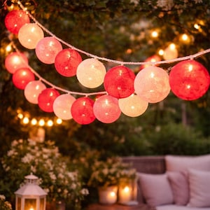 May include: A decorative string of illuminated, round lights in red and pink hues. The lights are strung on a white cord and are hanging outdoors. The background is blurred, with greenery and other lights visible.