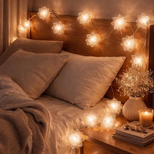 May include: A bedroom scene featuring a wooden headboard, pillows, and a bedspread. A string of illuminated, flower-shaped lights is draped across the headboard. A vase of flowers, a candle, and books sit on the nightstand, creating a cozy ambiance.