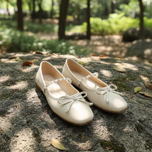 Puede incluir: Un par de bailarinas de color marfil con punta redonda y un pequeño lazo. Los zapatos tienen una correa adornada con pequeñas cuentas brillantes. El fondo es un entorno natural al aire libre con árboles y una superficie de piedra.