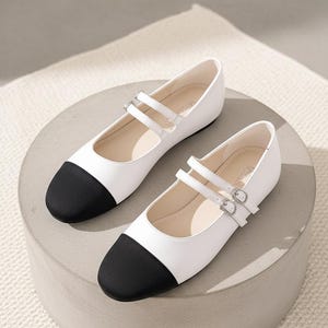 Puede incluir: Un par de bailarinas blancas y negras estilo Mary Jane con doble correa y cierres de hebilla. Los zapatos tienen una puntera redondeada con una puntera negra y se exhiben sobre un pedestal circular gris.