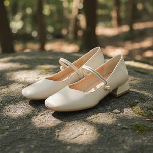 Puede incluir: Un par de zapatos Mary Jane color crema, con punta cuadrada y tacón de bloque pequeño. Los zapatos tienen una correa en la parte superior con un detalle de perla. Los zapatos están sobre una superficie de piedra. Los tacones miden unos 4 cm.