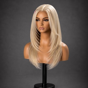 May include: A shoulder-length blonde wig with a center part and layered ends, displayed on a mannequin head. The hair is a light platinum shade with subtle darker roots, featuring a small pink decorative clip on the right side of the hairline.