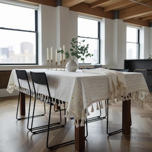 May include: A dining table set with a cream-colored tablecloth featuring yellow dot patterns and tassel trim. The table is set with black chairs, gold candle holders, and a white vase with greenery. The room has large windows and hardwood floors.