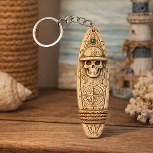 May include: A beige surfboard-shaped keyring featuring a skull in a hat, a compass design, and a green gem. The keyring is attached to a silver metal ring and chain. The design is carved into the surface.