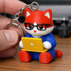 Op de afbeelding: Een rode, witte en blauwe katvormige sleutelhanger met een bril en een gele laptop. De kat draagt een blauw shirt en rode schoenen. De sleutelhanger is bevestigd aan een zilveren ketting.