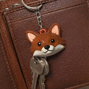 May include: A brown fox head keychain with a silver chain and keyring, holding two keys. The fox has a white muzzle, pink ears, and black eyes. The keychain is set against a brown leather wallet.