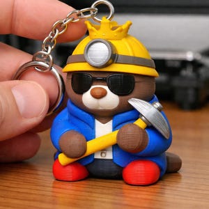 May include: A 3D-printed keychain featuring a cartoon bear character. The bear wears a yellow hard hat with a silver headlamp, black sunglasses, and a blue jacket. It holds a yellow pickaxe with a silver head and has red shoes.