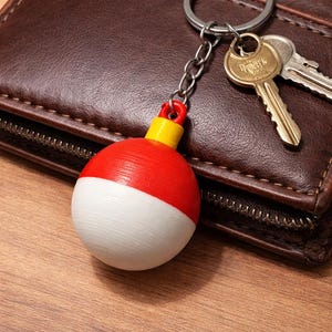 Könnte beinhalten: Ein roter, weißer und gelber Angelposen-Schlüsselanhänger mit einer silbernen Kette. Der Schlüsselanhänger befindet sich neben einer braunen Ledergeldbörse und zwei Schlüsseln auf einer Holzoberfläche.