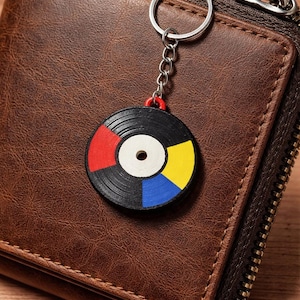 Könnte beinhalten: Ein schwarzer Vinyl-Schlüsselanhänger mit roten, gelben und blauen Akzenten. Der Schlüsselanhänger ist an einer silbernen Kette und einem Ring befestigt und liegt auf einer braunen Ledergeldbörse mit Reißverschluss.