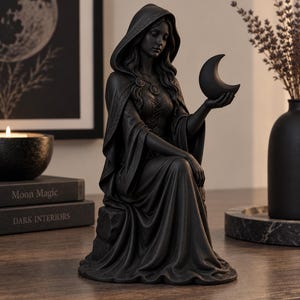 May include: A matte black figurine of a hooded figure sitting on a stone, wearing a long gown with a lace-up bodice and a cloak. The figure holds a crescent moon in one hand and wears a circular pendant necklace. The sculpture has intricate details.