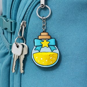 May include: A novelty keychain with a potion bottle design, filled with yellow liquid and a blue bow. The keychain is attached to a teal backpack, with keys hanging nearby. The bottle has a star in the center.