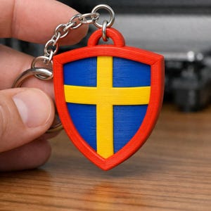 May include: A shield-shaped keychain featuring the flag of Sweden. The keychain has a red border, a blue center with a yellow cross, and a silver chain. The design is a patriotic accessory.