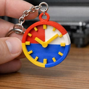 Puede incluir: Un llavero colorido en forma de reloj con un diseño rojo, blanco, azul y amarillo. La esfera del reloj tiene manecillas y marcadores amarillos. El llavero está sujeto a una cadena y un anillo plateados.