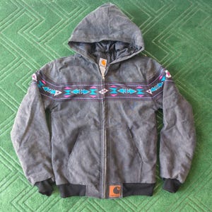 Può includere: Giacca grigia con cappuccio, cerniera a tutta lunghezza e orlo inferiore nero. La giacca presenta un colorato motivo geometrico sul petto e sulle maniche, con accenti rosa, blu e bianchi. Il logo Carhartt è visibile in basso a destra.