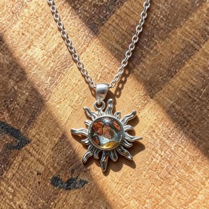 May include: A silver necklace featuring a sun pendant with a detailed design. The pendant has a central, colorful stone and is suspended from a delicate chain. The necklace is displayed on a round, wooden surface with a natural grain.