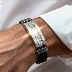 Puede incluir: Una pulsera plateada y negra con un diseño central texturizado se muestra en una muñeca. La pulsera tiene una pieza central rectangular plateada con una superficie estampada y una correa de cuero negro. La persona lleva una camisa blanca.