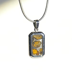 Può includere: Una collana color argento con un ciondolo rettangolare dotato di una cornice in metallo lucido. Il centro del ciondolo contiene tre pietre incastonate, irregolari e color ambra, su una base metallica rettangolare liscia.