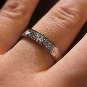 May include: A silver-toned metal ring featuring a flat band with a distinct cross-hatched, etched pattern across its surface. The band has a smooth, polished edge on both the top and bottom, creating a subtle contrast with the textured center.