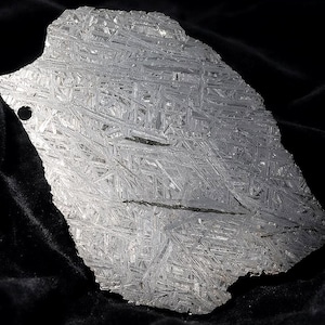 May include: A flat, metallic meteorite slice featuring a distinct Widmanstätten pattern of intersecting crystalline lines across its silver-grey surface. The irregular, jagged-edged specimen includes a small, circular hole near the upper left corner.