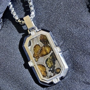 Pode incluir: Um colar com pendente retangular apresentando uma moldura de metal prateado polido com quatro pequenas pedras transparentes incrustadas. O centro exibe um padrão manchado de pedra translúcida em tons de âmbar e castanho escuro. Com corrente.