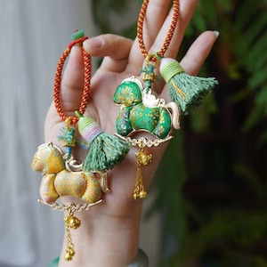 Pode incluir: Dois pingentes decorativos de cavalo com detalhes dourados e borlas. Um cavalo é amarelo e o outro verde, ambos com intrincados padrões florais. Os pingentes pendem de cordões trançados vermelhos e dourados.