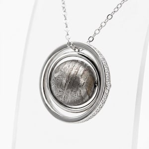 Puede incluir: Un collar con colgante de tono plateado que presenta una cuenta central esférica de meteorito con un patrón de Widmanstätten. La esfera está encerrada en dos anillos concéntricos de plata, con el anillo exterior decorado con pequeñas piedras claras.