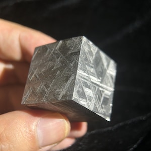 Peut inclure: Un cube métallique présentant un motif de Widmanstätten distinct composé de lignes entrecroisées sur ses surfaces grises polies. L'objet affiche une structure cristalline aux nuances d'argent et de charbon, reflétant la lumière sur ses faces.