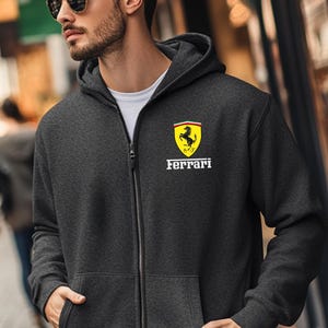 May include: Charcoal gray zip-up hoodie with the Ferrari logo on the chest. The logo features a black prancing horse on a yellow background, with the word "Ferrari" in white below. The hoodie has a drawstring hood and a white undershirt is visible.