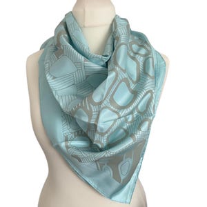May include: A light blue silk scarf with a geometric pattern in shades of gray. The scarf is draped over a mannequin, showcasing its square shape and smooth texture. The design features organic shapes and linear elements.