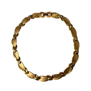 May include: A gold-toned necklace with a textured, leaf-shaped design. The necklace is composed of interconnected links, creating a continuous loop. The clasp is visible at the top. The jewelry is set against a white background.