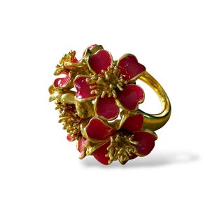 Op de afbeelding: Een goudkleurige ring met een cluster van rode en gouden bloemmotieven. De bloemen hebben rode bloemblaadjes en gouden centra, wat een levendig contrast creëert. De band is goudkleurig.