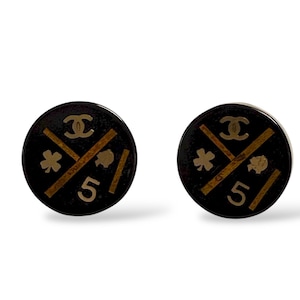 May include: Pair of black and gold round earrings. Each earring features a gold logo, crossed lines, a clover, a spade, and the number 5. The earrings have a glossy finish and a classic design.