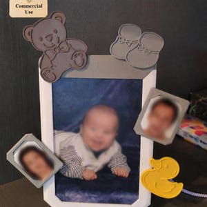Könnte beinhalten: Ein weißer Bilderrahmen mit einem Babyfoto, umgeben von grauen Magneten in Form eines Teddybären, Babyschuhen und einer gelben Gummiente. Zusätzliche kleine Fotorahmen sind ebenfalls angebracht. Das Bild hat einen blauen Hintergrund.