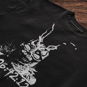 Op de afbeelding: Een zwart t-shirt met een witte grafische print van een konijnachtig wezen met lange oren en de tekst 28:06:42:12 in een verweerd stencil-lettertype op de voorzijde, voorzien van een ronde halslijn.