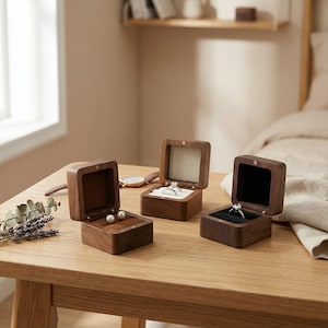 Könnte beinhalten: Drei offene Holzringboxen auf einem Holztisch, mit einer Uhr und Lavendelzweigen. Die Boxen zeigen einen Ring und zwei Perlenohrringe. Die Boxen sind aus dunklem Holz mit einem Samtinnenraum.