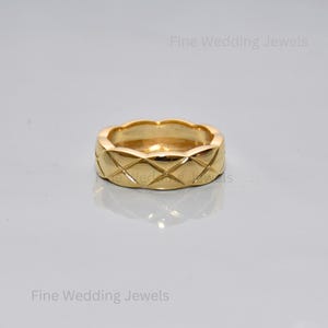 May include: A gold-colored ring with a quilted design. The ring has a rounded shape with a series of diamond-shaped patterns across its surface. The ring is a wedding band.