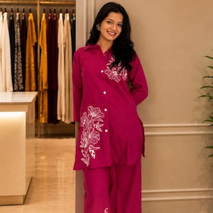 Peut inclure: Une femme porte un ensemble deux pièces magenta avec des broderies florales blanches. L'ensemble comprend une chemise boutonnée et un pantalon large. En arrière-plan, un intérieur de magasin avec des présentoirs de vêtements et l'enseigne "Lavish Enterprises".