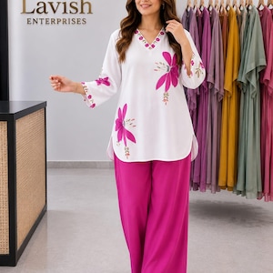 May include: A woman wearing a white tunic and fuchsia palazzo pants. The tunic has a V-neck and is adorned with large, pink floral designs. The pants are wide-legged. A rack of colorful clothing hangs in the background.