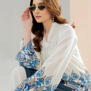 May include: A woman wearing a white blouse with blue and white patterned sleeves and a matching skirt. She is wearing gold-rimmed sunglasses and earrings. The blouse has a V-neck and the sleeves have a detailed paisley design.