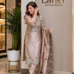 May include: A beige silk salwar kameez set with floral embroidery. The outfit includes a long tunic, fitted trousers, and a matching scarf. The embroidery is in shades of grey and black. The text "Lavish Enterprises" is visible.